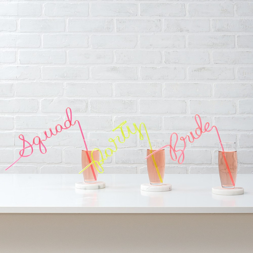 Three cocktail glasses with pink and yellow silly straws spelling Squad, Party, and Bride