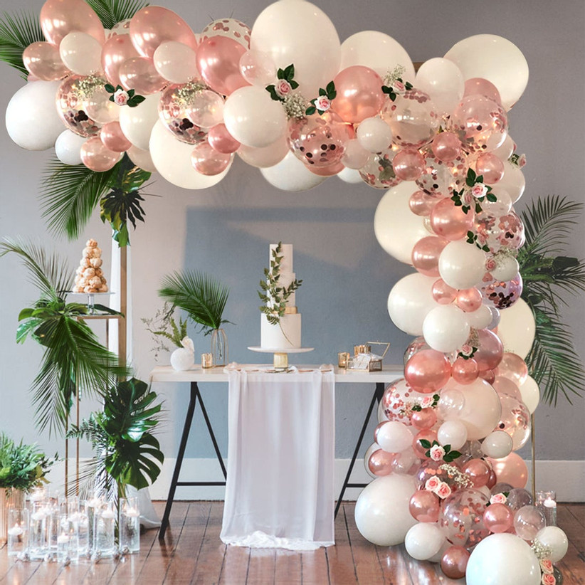 Extravagant rose gold and white balloon arch over cake table with tropical greenery