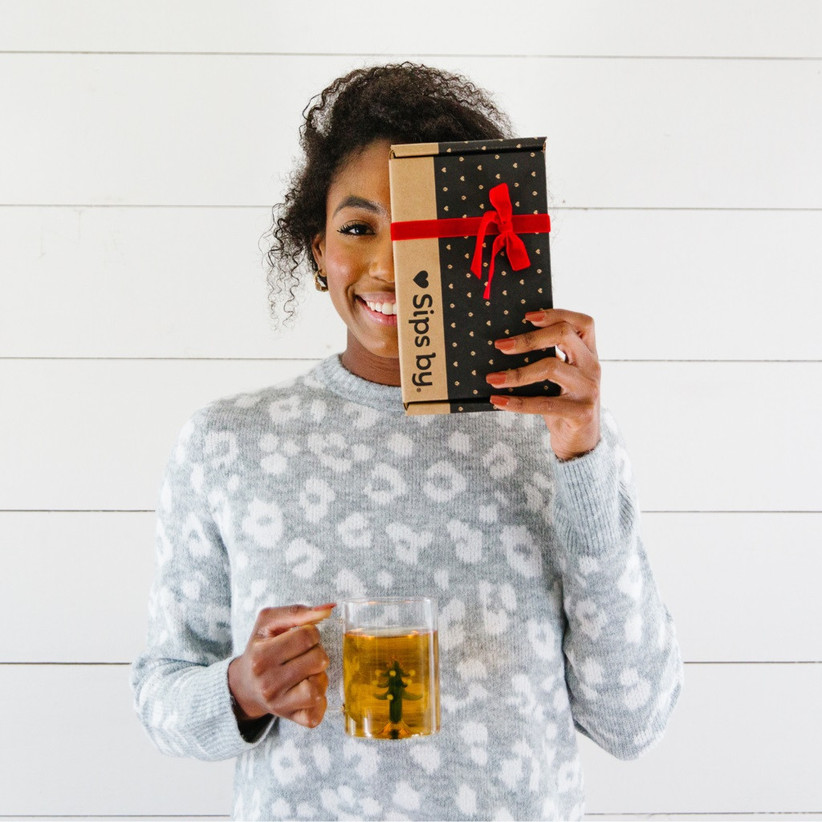 Woman smiling holding a glass mug of tea in one hand a Sips by subscription box in the other