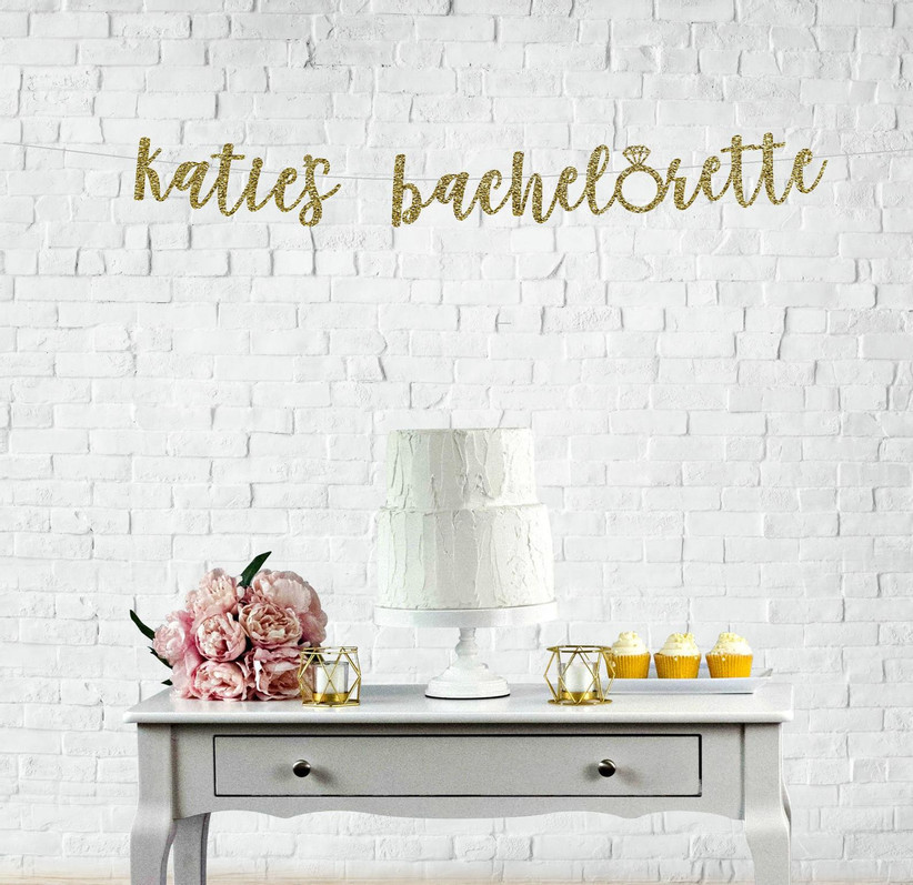 Personalized glittery gold bachelorette banner reading Katie's Bachelorette displayed over a vintage cake table