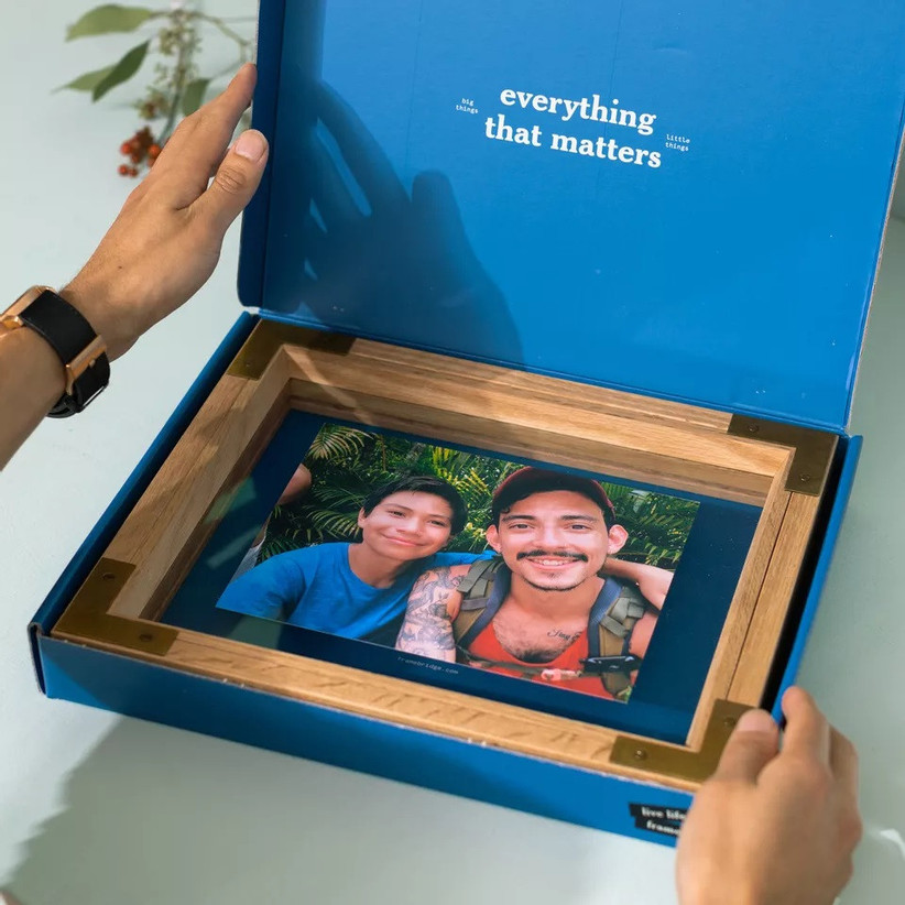 Person holding blue gift box open with wooden photo frame inside depicting two people