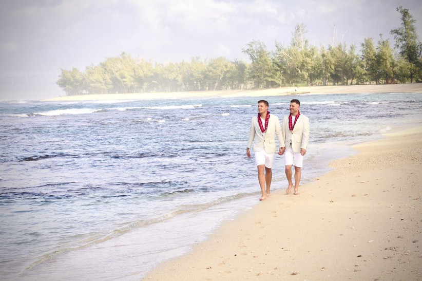 ЛГБТ + пара двух женихов гуляет по пляжу в шортах и ​​розовых цветочных леях в тон