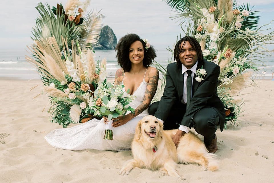 A newlywed couple posing with their dog in front of a beach wedding ceremony arch at Pelican Pub & Brewery